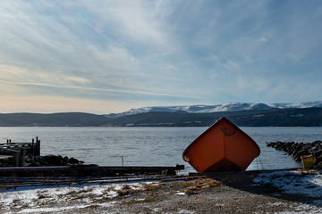 An orange wooden boat pulled up on a slipway near the ocean with snow covered mountains in the background. The sky is blue and cloudy. The evening sky has a tinge pink to the horizon.