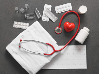 Stethoscope, doctor's uniform, cardiogram, heart and pills on grey background. Cardiology concept