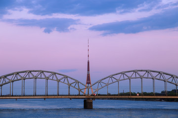 Vintage through (tied) arch railway bridge (Dzelzcela tilts) over Daugava river and the radio and TV tower (televizijas tornis) in Riga, Latvia at dawn, after sunset, with blue clouds in a pink sky.
