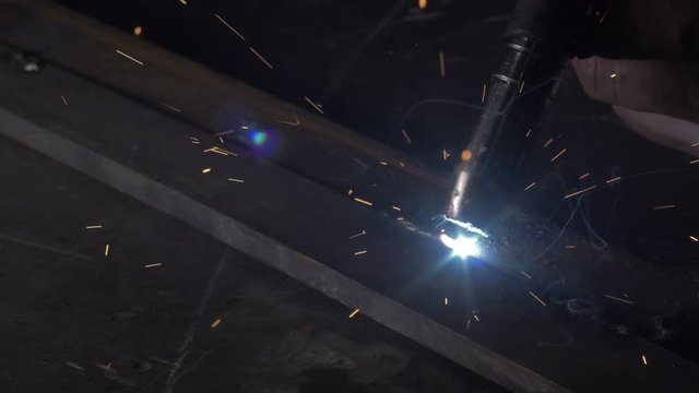 Forge workshop. Smithy. Worker in a welding hood helmet welds a part by electric welding. Sparks are reflected in the protective screen. Blacksmith makes iron product for manufacture. Slow motion.