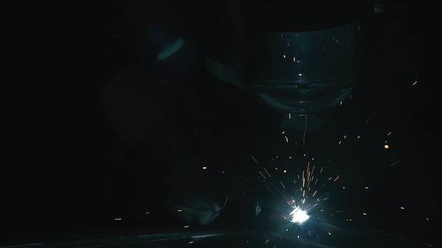 Forge workshop. Smithy. Worker in a welding hood helmet welds a part by electric welding. Sparks are reflected in the protective screen. Blacksmith makes iron product for manufacture. Slow motion.