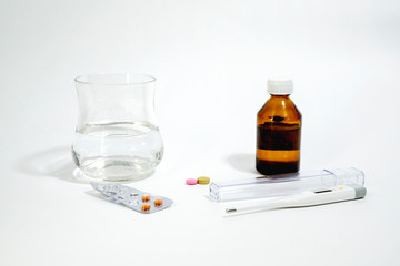 A glass of clean water on a white background, next to an electronic thermometer, pills and a package of pills.  Selective focus.
