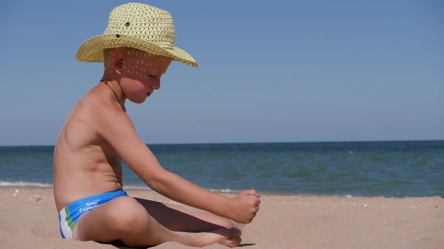 Boy sits on the Golden sand. The child wears a straw hat with a large brim. Child playing in the sand on the beach. Light green hat develops in the wind.