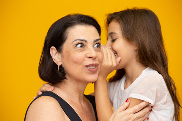 Mom and daughter secretive. kid sharing a secret with mother. Mommy little kid daughter isolated on yellow background studio portrait. Mother's Day love family parenthood childhood concept