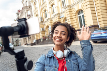 Outdoors Leisure. Young girl blogger with headphones walking holding stabilizer recording vlog on smartphone talking playful close-up