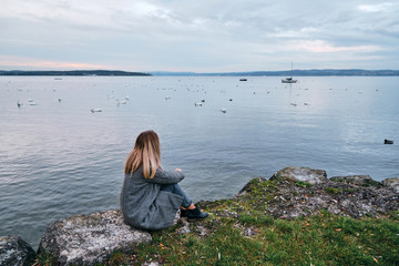 Naklejka premium Beautiful blond hair woman enjoy panorama by the Bodensee lake. Landscape in Switzerland. Happy girl in travel. Amazing scenic outdoors view. Dramatic sky. Swans on water. Adventure lifestyle, freedom