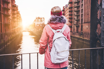 Rear view of adult woman tourist with white backpack enjoying sunset on the bridge in Speicherstadt historical warehouse district. Hamburg, Germany, Europe
