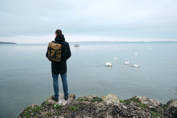 Fototapeta na wymiar Man with backpack enjoy panorama by the Bodensee lake. Landscape in Switzerland. Happy man in travel. Amazing scenic outdoors view. Dramatic sky. Swans on water. Adventure lifestyle, freedom