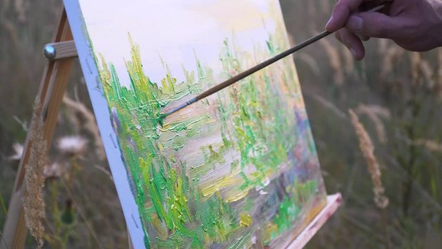 Male artist paints sunset landscape in open space amidst natural landscape. Close-up view of canvas and brush movements in a hand