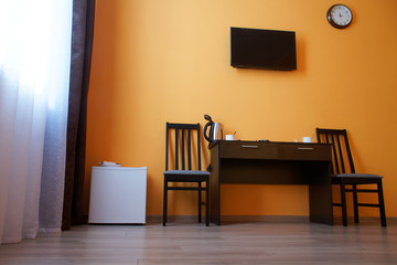 Compact white refrigerator, a table, two chairs, an electric kettle and a wall-mounted TV in the interior of a small studio apartment or hotel room
