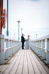 The bride and groom walk along the painted light wooden pier on a cloudy summer day, holding hands. The bride is pregnant. Helsinki. Finland