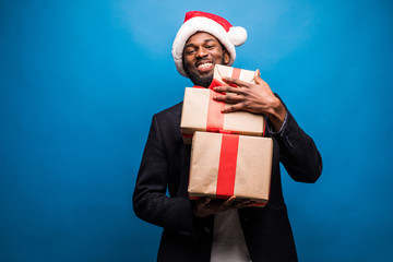 Young African American man wearing a santa hat offering a gifts on blue background