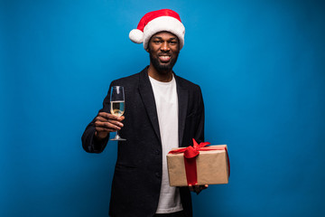 African young man wearing santa hat with champagne and gift box against blue background. Happy young black guy enjoying christmas eve.