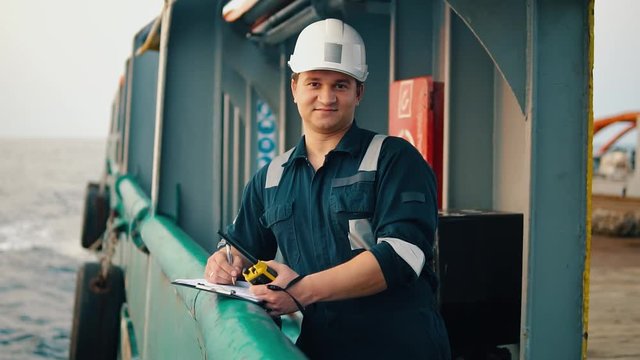 Marine chief officer or chief mate on deck of ship or vessel. He fills up ahts vessel checklist. Ship routine paperwork. He holds VHF walkie-talkie radio in hands.