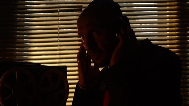 Secret agent is listening to and recording conversations, reel tape recorder, the light through the blinds, headphones on his head