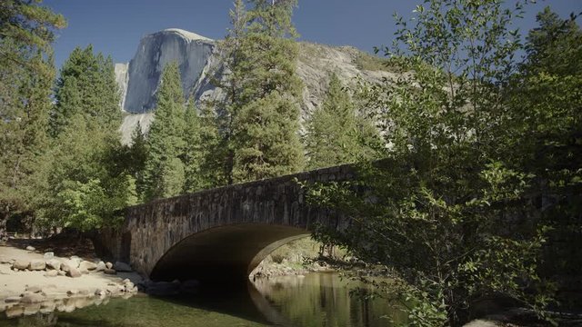 Tilt up to scenic view of Stoneman Bridge over river near trees and mountains / Yosemite Valley, California, United States