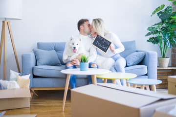 Young beautiful couple with dog sitting on the sofa holding blackboard with message kissing at new home around cardboard boxes