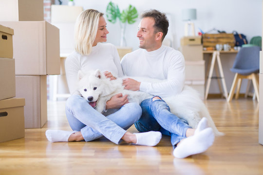 Young beautiful couple with dog sitting on the floor at new home around cardboard boxes