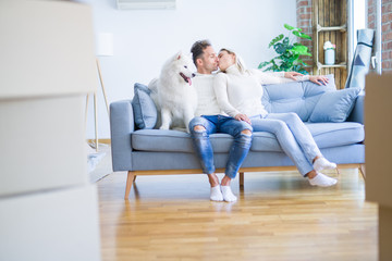 Young beautiful couple with dog kissing sitting on the sofa at new home around cardboard boxes