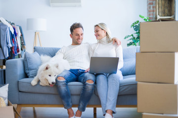 Fototapeta premium Young beautiful couple with dog sitting on the sofa using laptop at new home around cardboard boxes