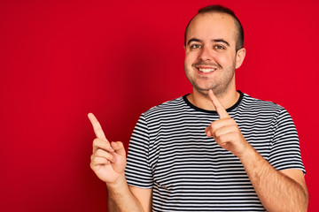 Fototapeta premium Young man wearing striped navy t-shirt standing over isolated red background smiling and looking at the camera pointing with two hands and fingers to the side.