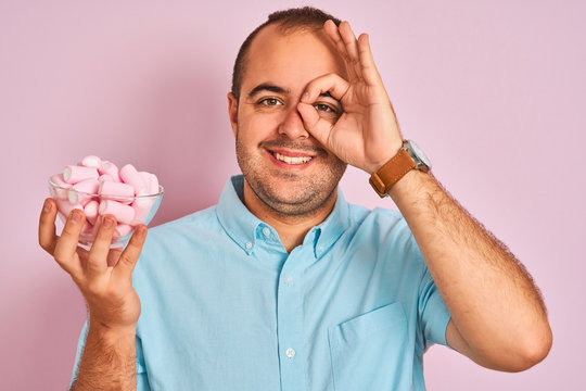 Young man holding bowl with marshmallows standing over isolated pink background with happy face smiling doing ok sign with hand on eye looking through fingers