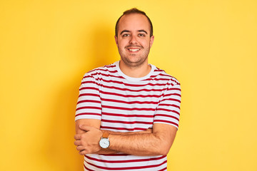 Young man wearing casual striped t-shirt standing over isolated yellow background happy face smiling with crossed arms looking at the camera. Positive person. - Powered by Adobe