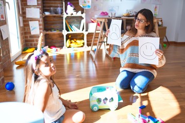 Beautiful pshychologist and toddler doing therapy with emotions emoji around lots of toys at kindergarten