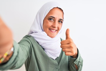 Young Arab woman wearing hijab taking selfie photo over isolated background happy with big smile doing ok sign, thumb up with fingers, excellent sign