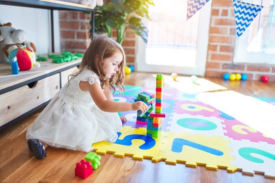 Adorable blonde toddler playing with building blocks toy around lots of toys at kindergarten