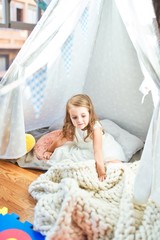 Adorable blonde toddler playing sitting on blanket inside tipi at kindergarten