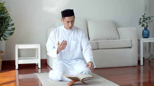 Young Muslim man wearing Islamic clothes reading Quran and praying