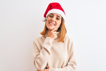 Beautiful redhead woman wearing christmas hat over isolated background looking confident at the camera smiling with crossed arms and hand raised on chin. Thinking positive.