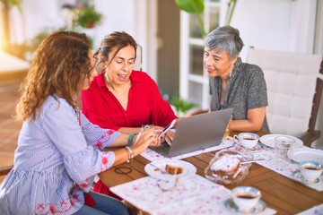 Obraz premium Meeting of middle age women having lunch and drinking coffee. Mature friends smiling happy using laptop at home on a sunny day