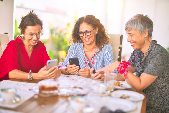 Meeting of middle age women having lunch and drinking coffee. Mature friends smiling happy using smartphone at home on a sunny day