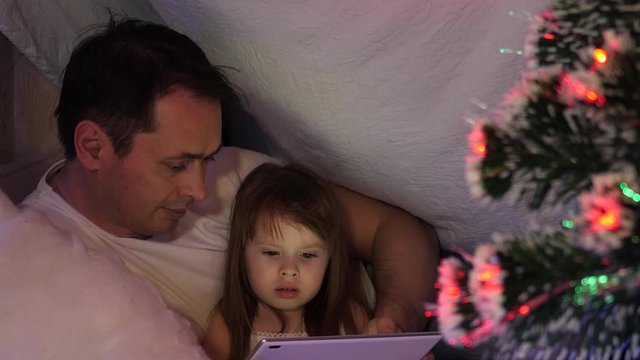 dad and daughter on Christmas evening, play and watch cartoons on tablet, in a children's room in tent with garlands. baby and Father are playing in room. concept of happy childhood and family.
