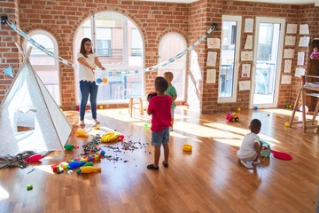 Young beautiful teacher and toddlers playing basketball around lots of toys at kindergarten