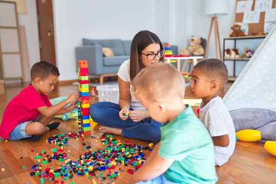 Young beautiful teacher and toddlers playing with building blocks around lots of toys at kindergarten