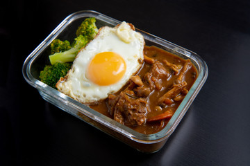 Beef curry rice in glass lunch box on black wooden dining table ( Onsen tamago, Potato, enoki mushroom, onion, broccoli, Japanese style)
