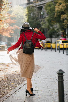 Happy slim girl in black high heel shoes dancing in park in autumn day. Outdoor full-length photo from back of brunette graceful woman in hat.