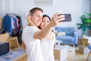 Young beautiful couple standing using smartphone to take a selfie at new home around cardboard boxes