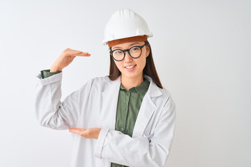 Young chinese engineer woman wearing coat helmet glasses over isolated white background gesturing with hands showing big and large size sign, measure symbol. Smiling looking at the camera. Measuring