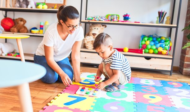 Beautiful teacher and toddler boy sitting on puzzle playing with numbers at kindergarten