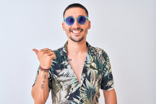 Young handsome man wearing Hawaiian sumer shirt and blue sunglasses over isolated background smiling with happy face looking and pointing to the side with thumb up.