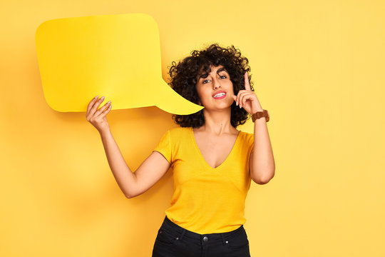 Young Arab Woman With Curly Hair Holding Speech Bubble Over Isolated Yellow Background Surprised With An Idea Or Question Pointing Finger With Happy Face, Number One