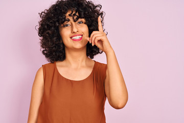 Young arab woman with curly hair wearing t-shirt over isolated pink background surprised with an idea or question pointing finger with happy face, number one