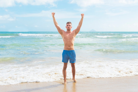 Handsome beautiful naked topless guy in swimming trunks, tourist enjoying vacation at sea, relaxing on beach. Young man standing with his hands up in air, raised arms, full length, celebrating success