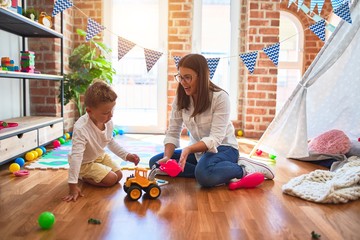 Beautiful teacher and toddler playing with tractor around lots of toys at kindergarten