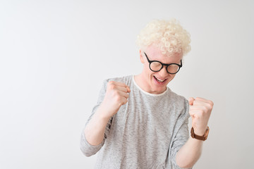Young albino blond man wearing striped t-shirt and glasses over isolated white background very happy and excited doing winner gesture with arms raised, smiling and screaming for success. Celebration 