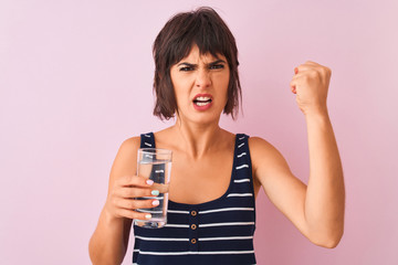 Young beautiful woman holding glass of water standing over isolated pink background annoyed and frustrated shouting with anger, crazy and yelling with raised hand, anger concept